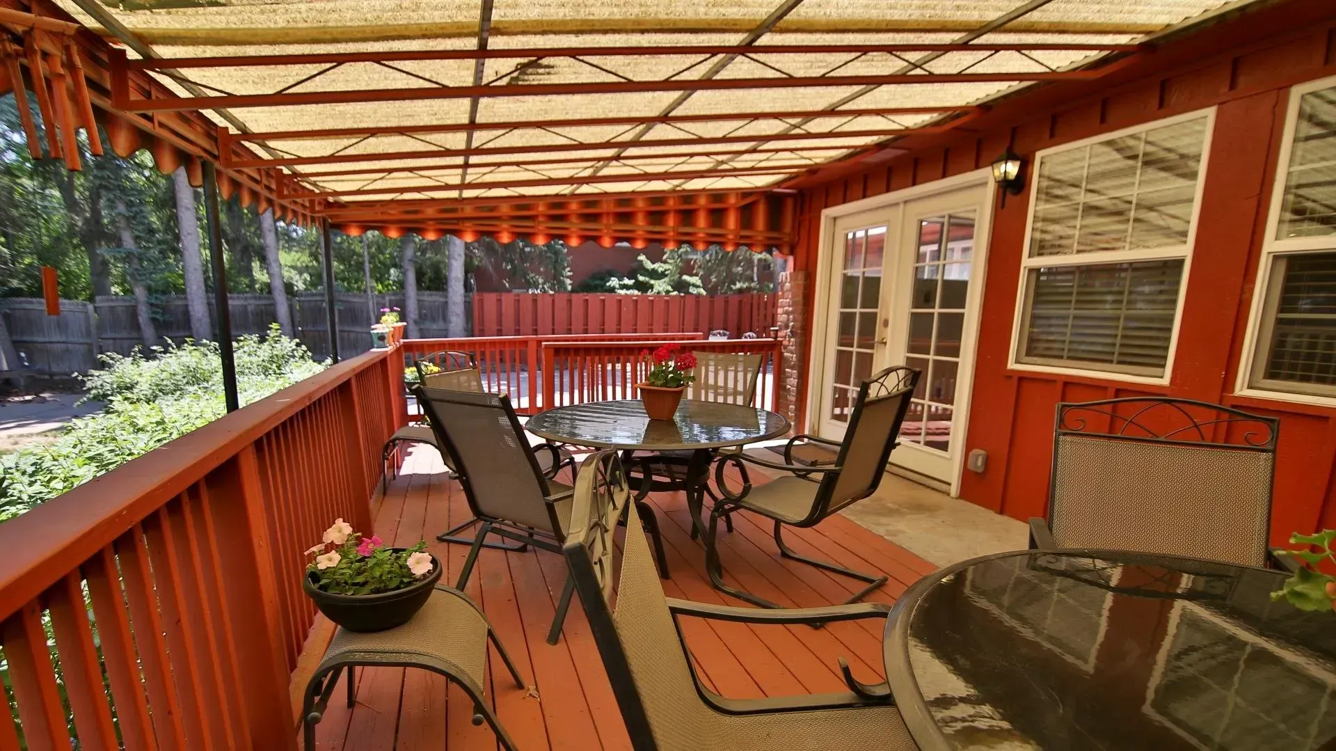 Red-painted deck with outdoor furniture, sheltered by a woven roof, with a view of a backyard.