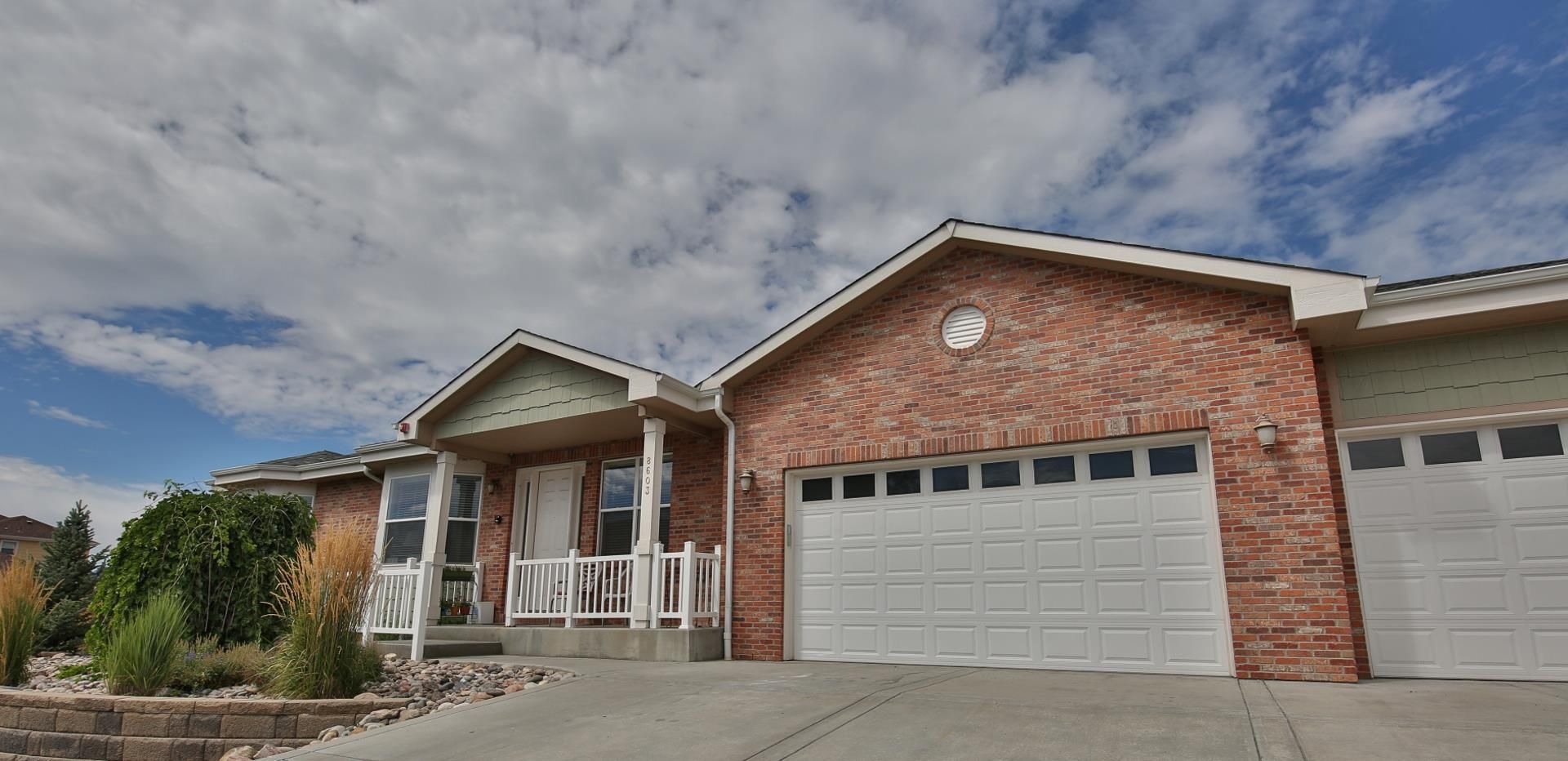 A brick house with a white garage door, a porch, and a cloudy blue sky above.