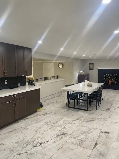 A modern dining area with a large table, dark cabinets, marble floors, and a raised platform in a well-lit setting.