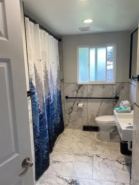 Accessible bathroom: white marble tile, blue shower curtain, toilet with grab bar, and small sink.