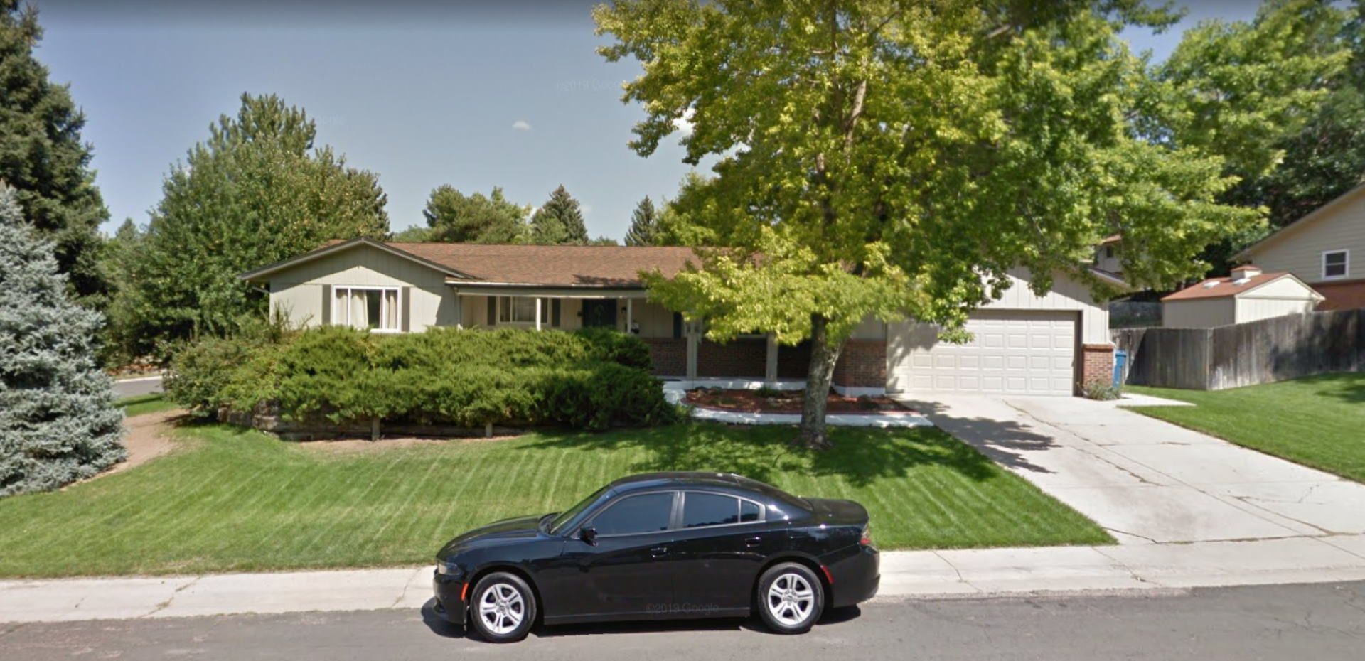 Black car parked in front of a beige house with brown roof, green lawn, and trees on a sunny day.
