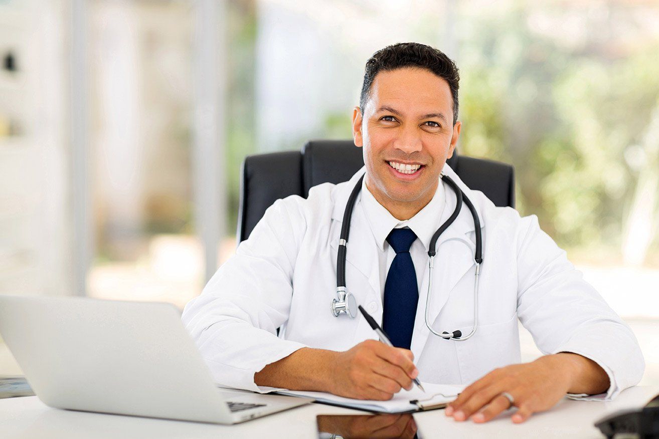 Doctor smiling at desk, wearing lab coat, stethoscope, and tie; writing on a clipboard in a bright office.