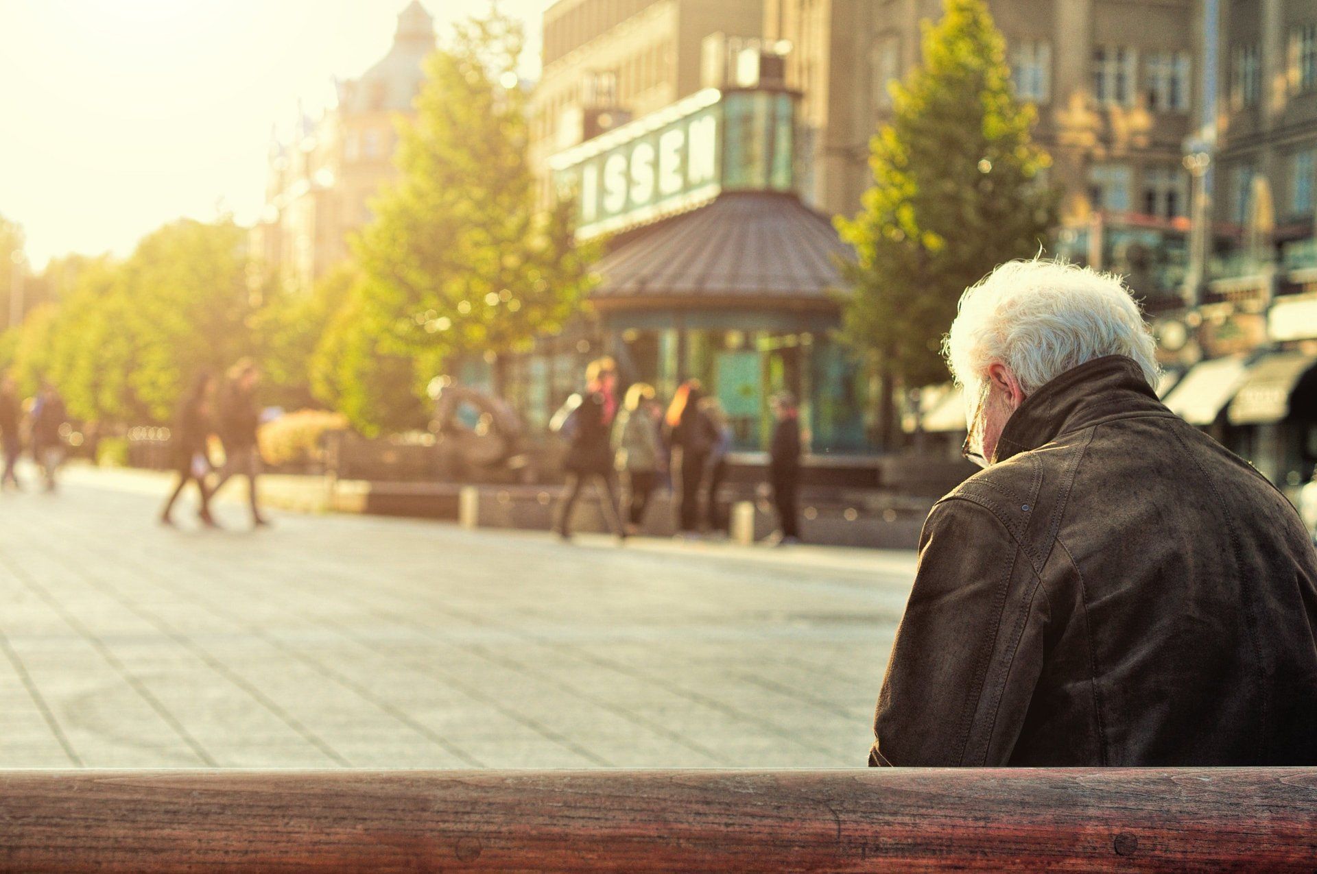 Elderly person with white hair sits, looking at a sunny city scene with blurred pedestrians and buildings.