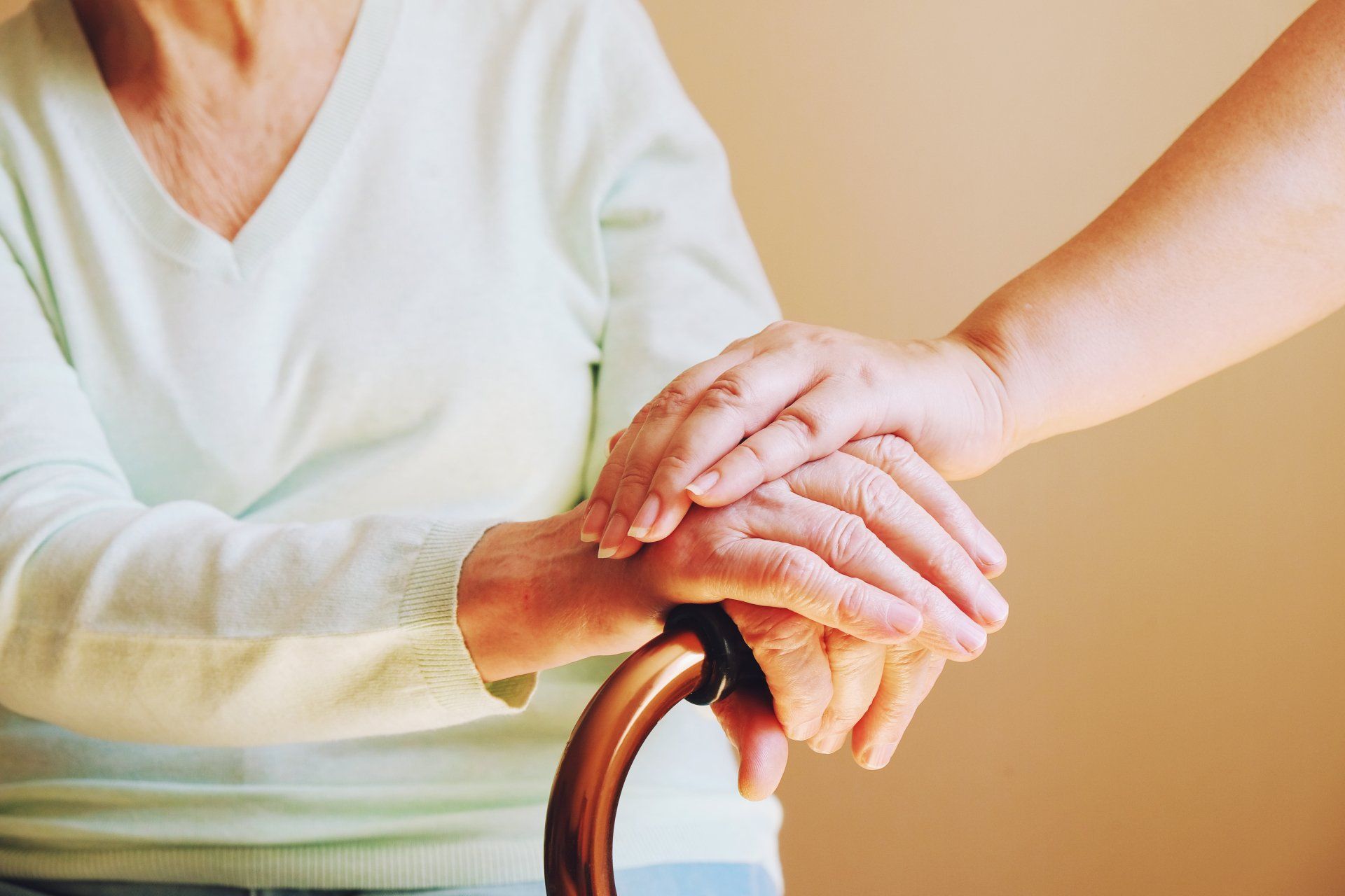 Person's hand resting on another's, over a cane handle. Light blue shirt, neutral background. Focus on care.