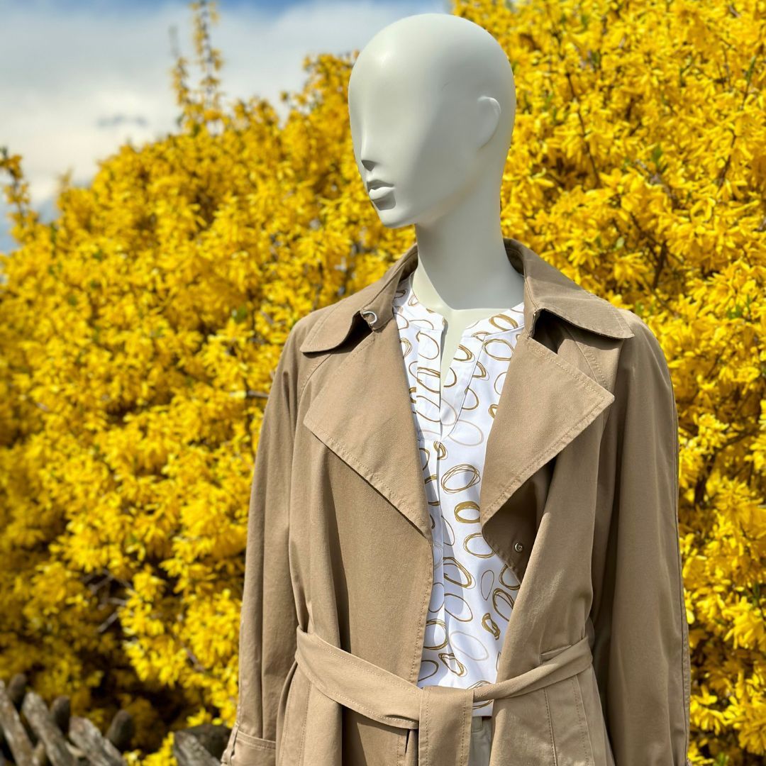 Eine Schaufensterpuppe mit einem Trenchcoat steht vor einem Busch mit gelben Blumen.