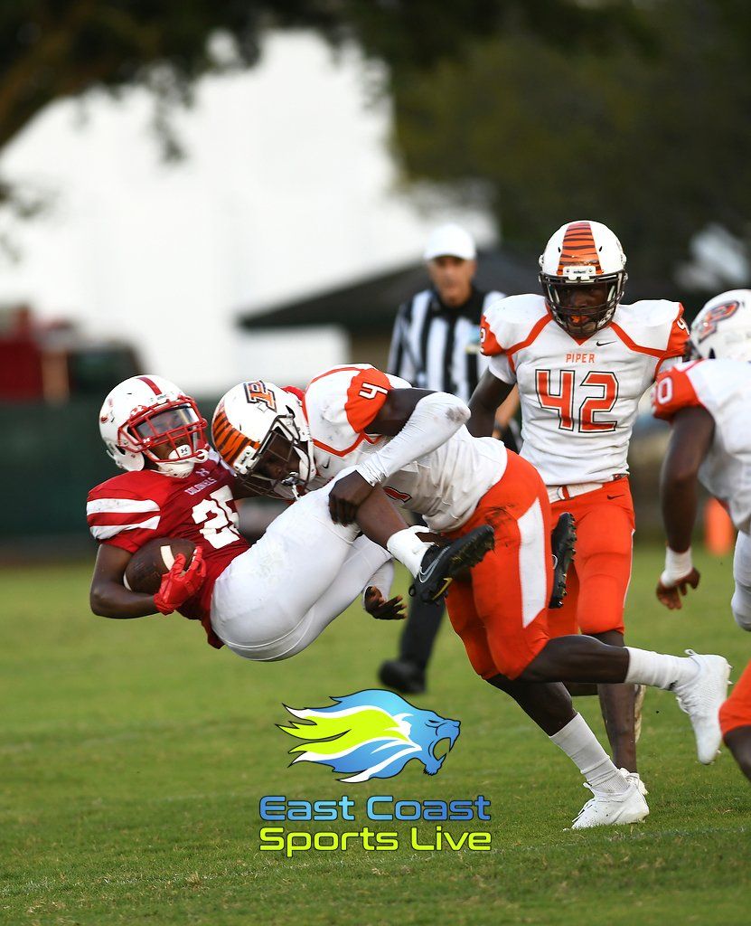 Football players in action; red and orange uniforms, tackling on a green field.