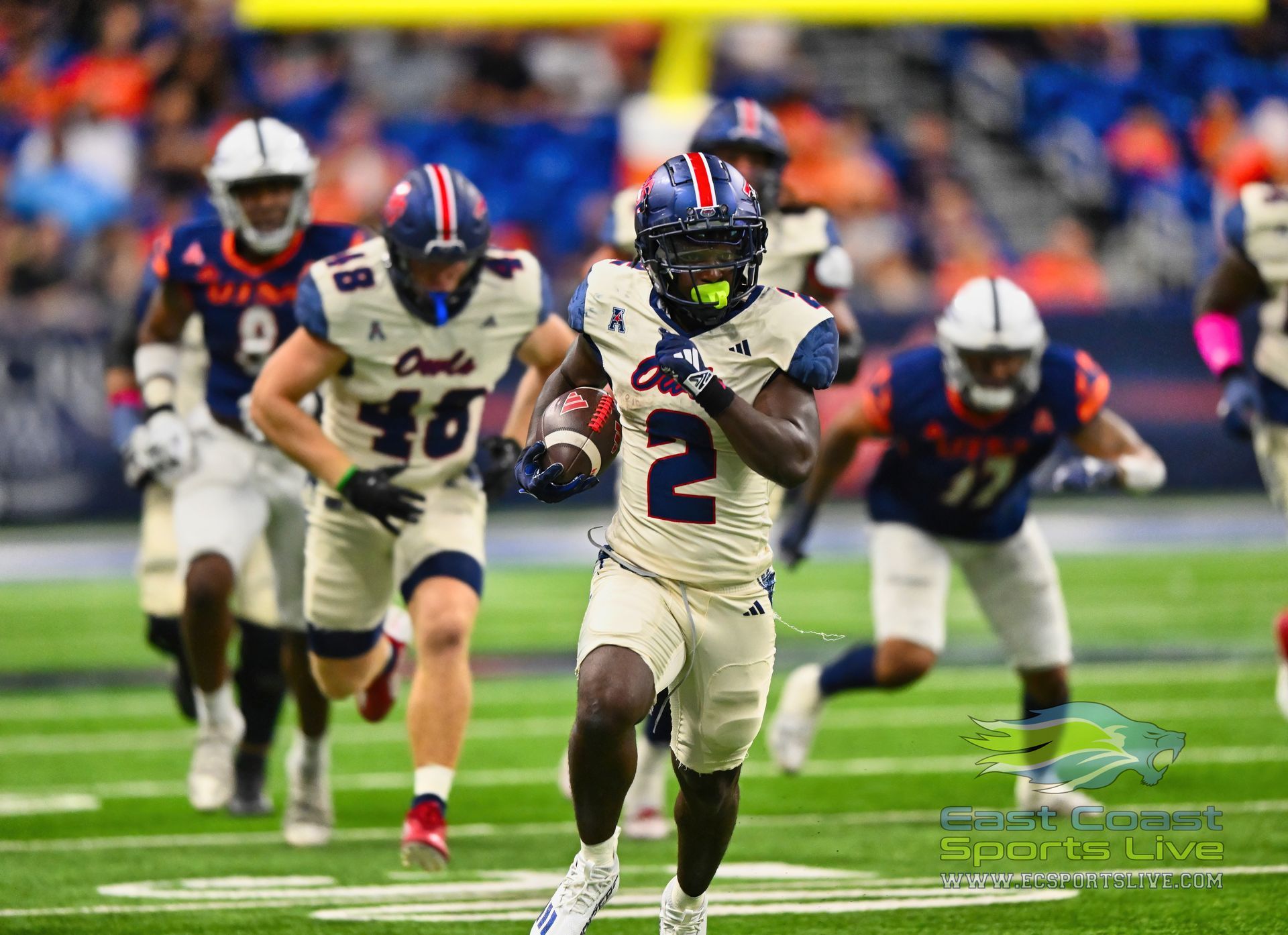 Football player, number 2, runs on a green field; white uniform with blue and red accents, blue sky background.