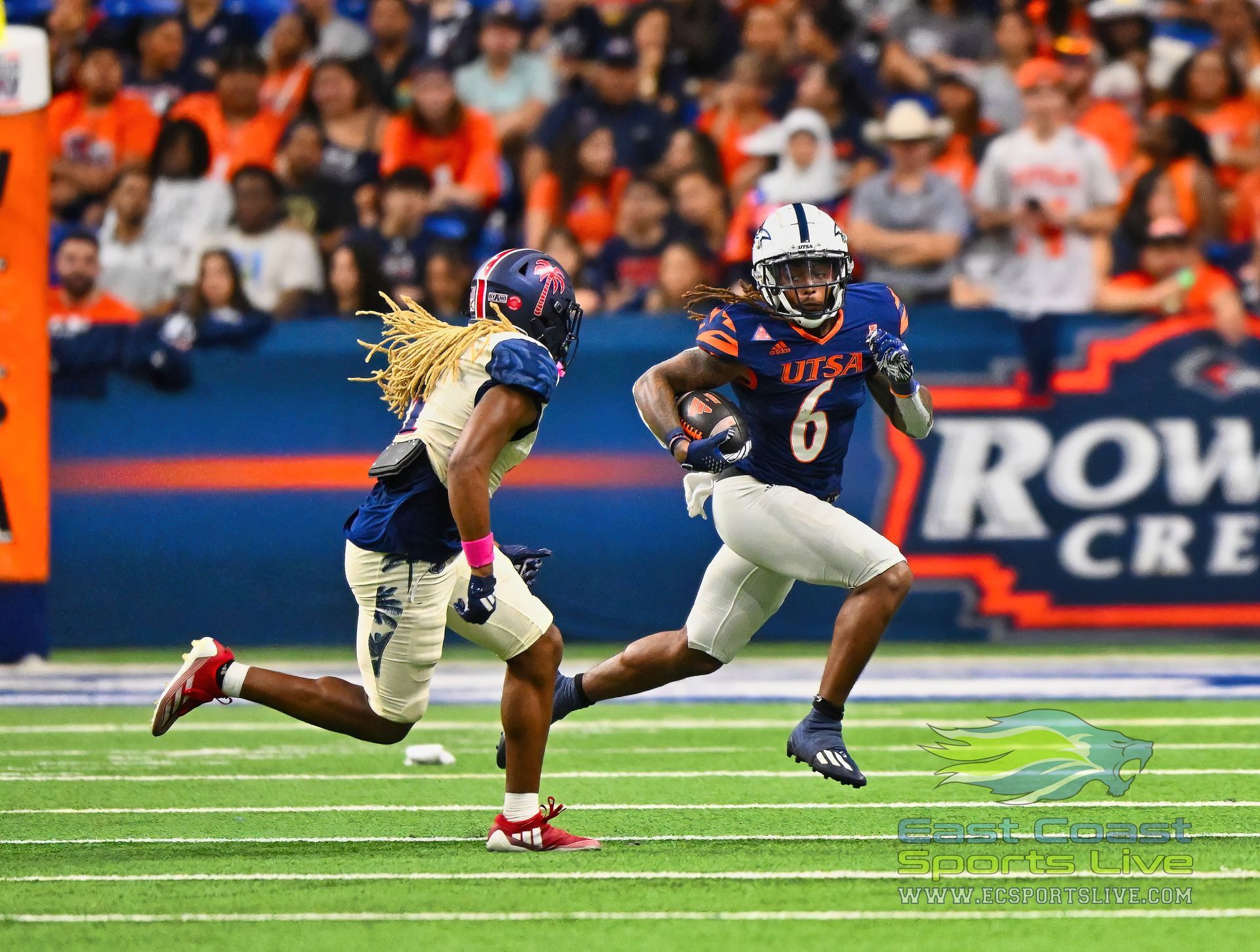 Football player in blue uniform runs with the ball, being pursued by another player. Stadium crowd in background.