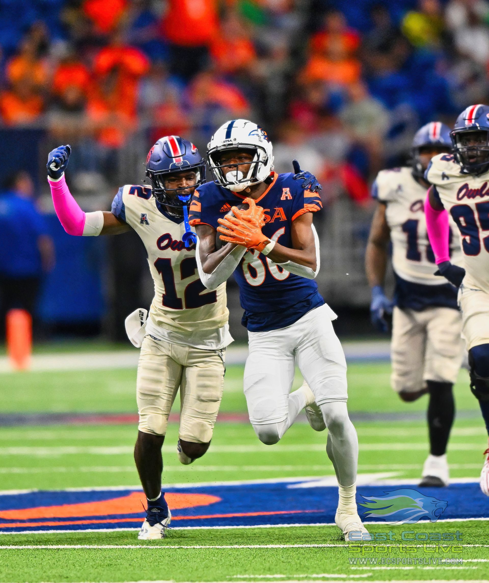 Football players in blue and orange uniforms on a green field. One player with ball, another attempting to tackle.