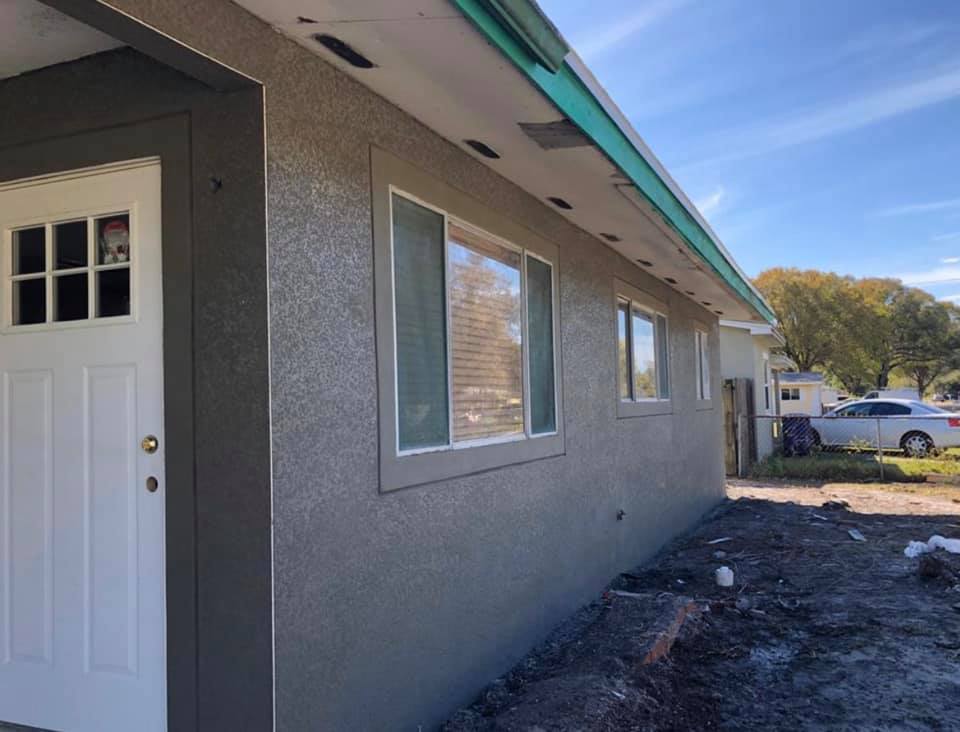 Exterior of a house with gray stucco walls, white door, and windows. Blue sky, fall trees in the background.