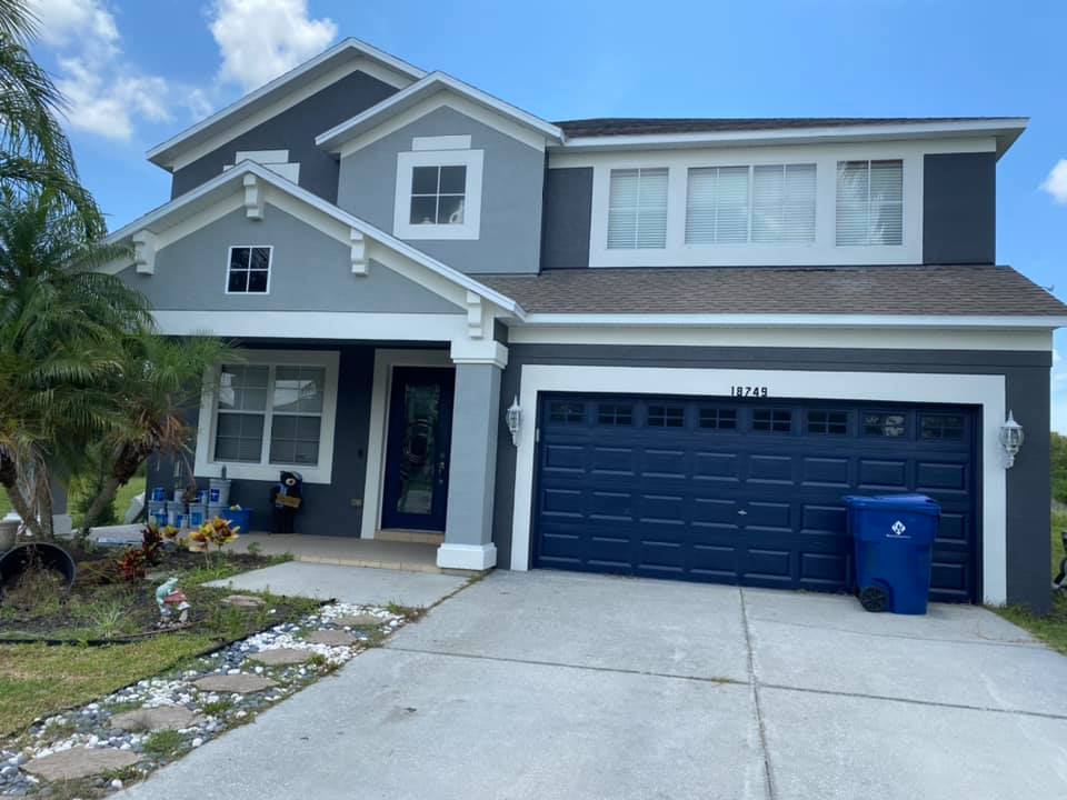 Two-story house with gray exterior, blue garage door, and blue trash can on a concrete driveway under a blue sky.