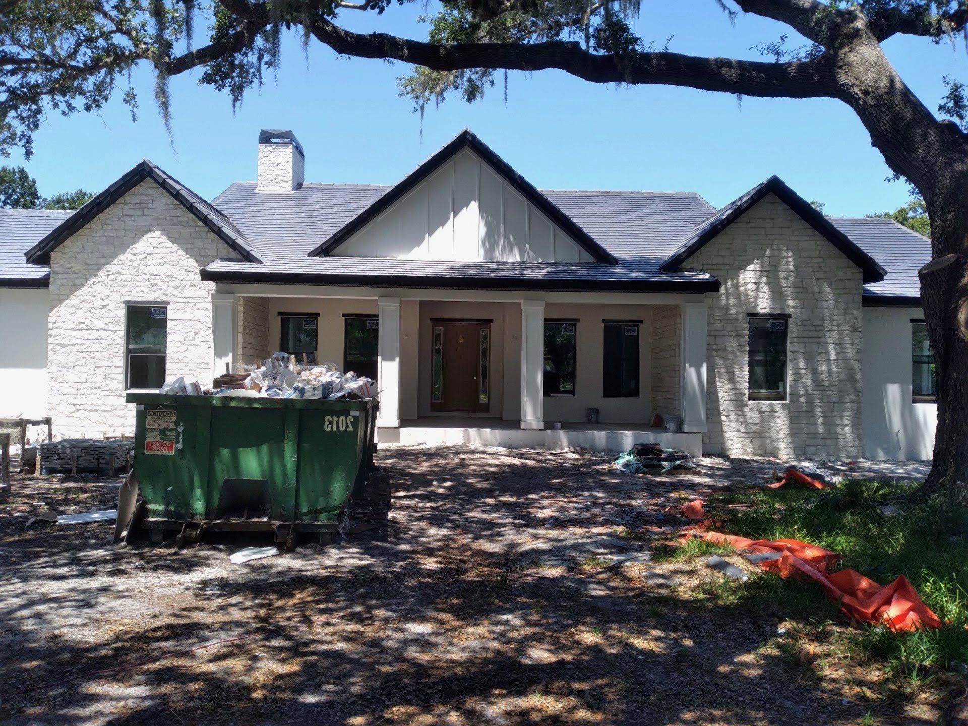A house under construction with a green dumpster in front. White exterior with black trim, blue roof.
