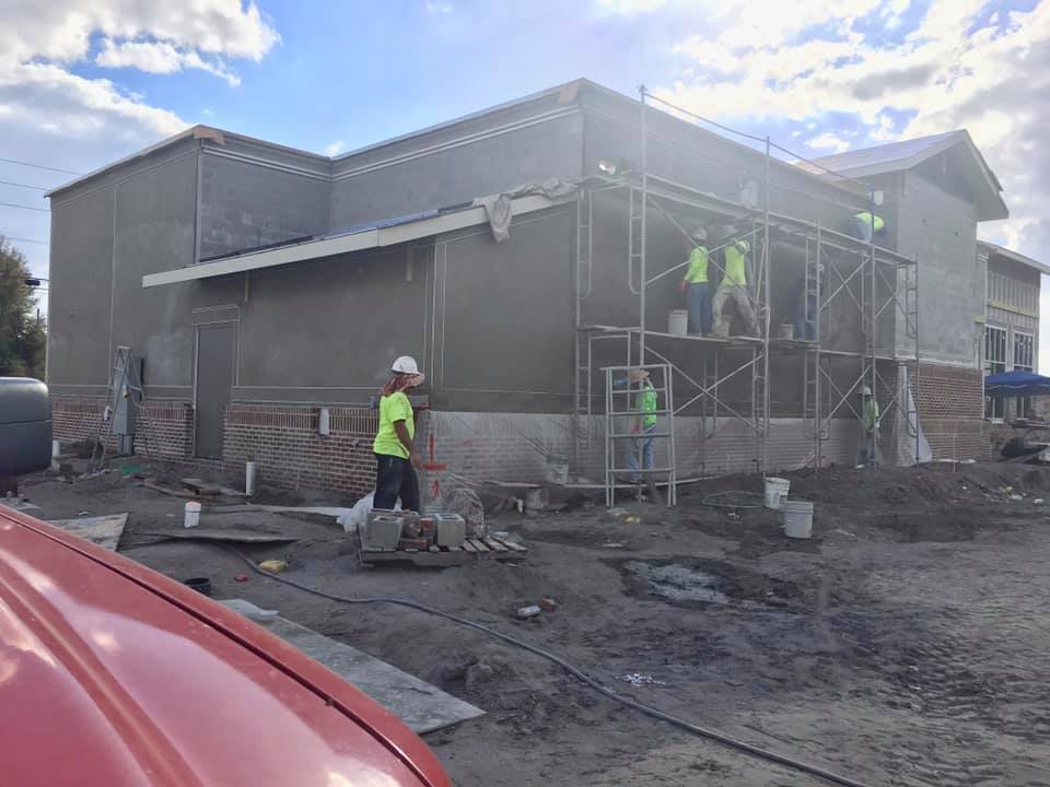 Construction workers plastering exterior walls of a building; brick facade at the bottom, scaffolding, and a red vehicle in the foreground.