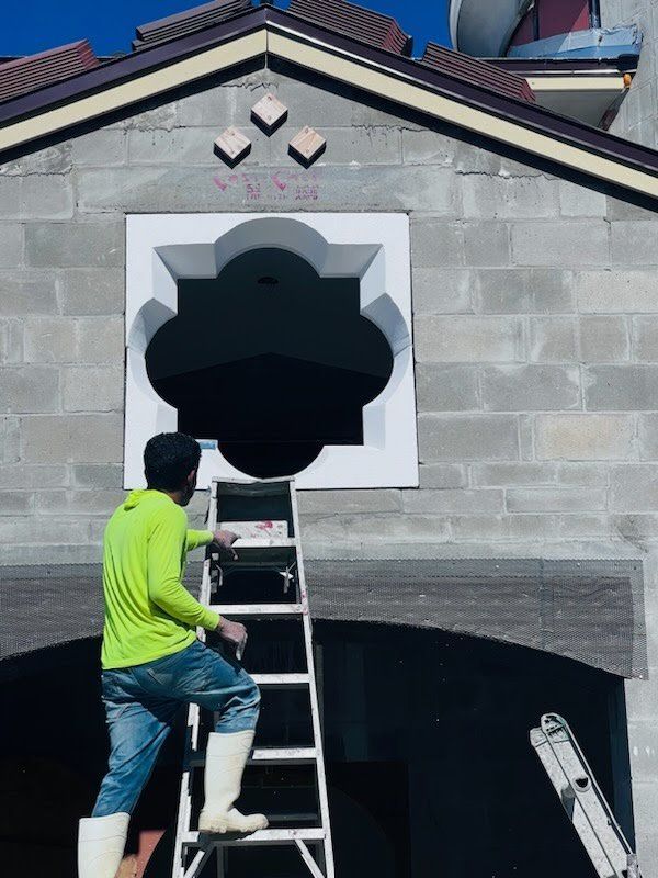 Construction worker on a ladder near a white ornate window. Building has gray brick, bright blue sky.