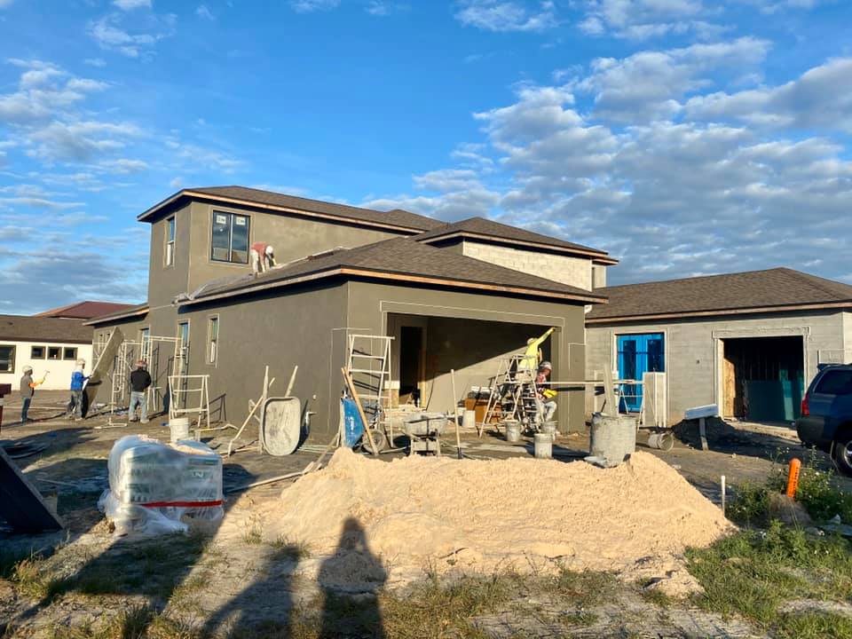 Construction of a two-story house with attached garage under a partly cloudy blue sky; workers are present.