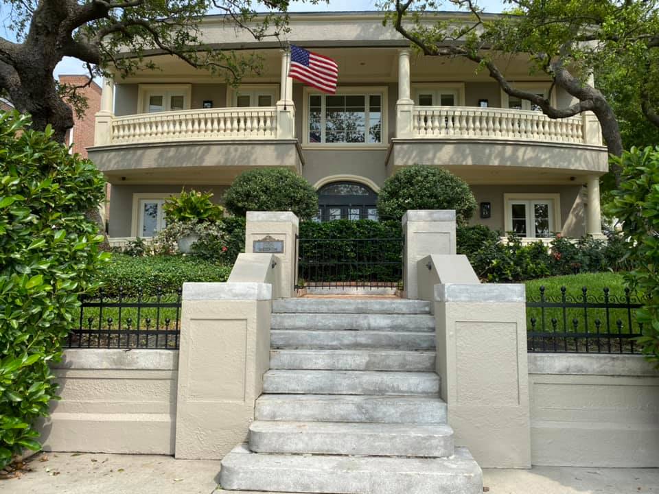 Two-story beige house with American flag, steps, and a black wrought-iron fence.