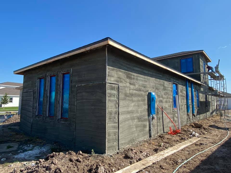 House under construction; exterior concrete walls; blue-covered windows; scaffolding; clear sky; dirt ground.