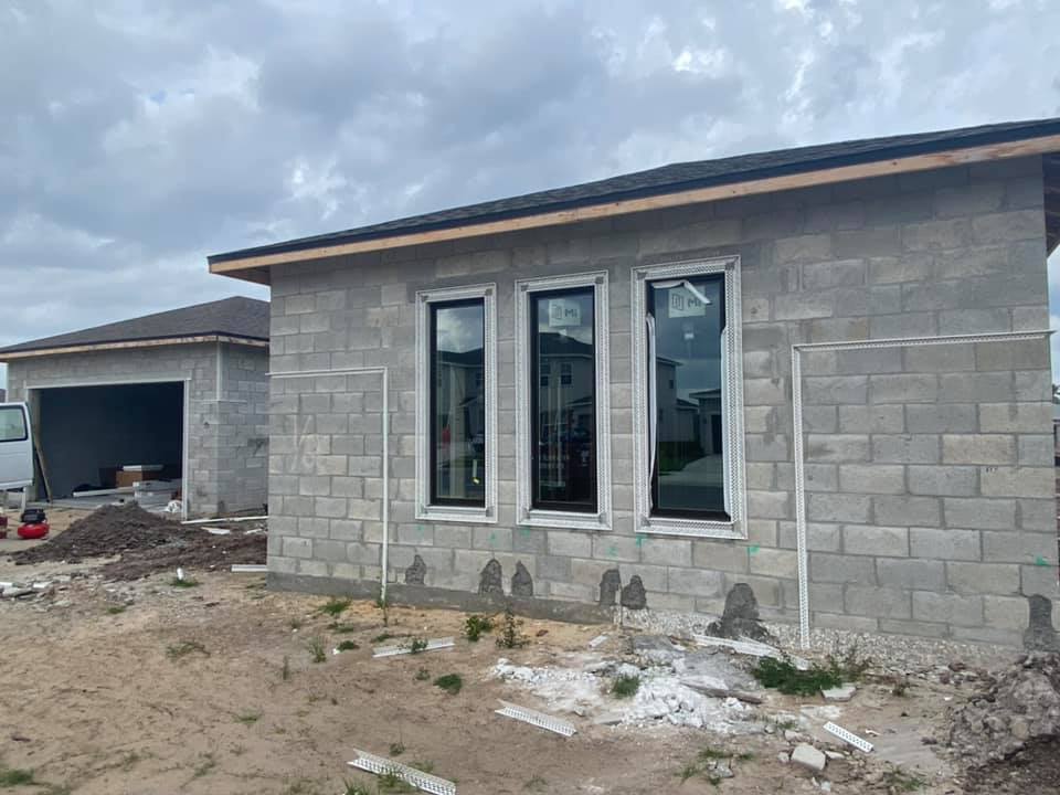 Construction site with cinder block house and garage. Three black-framed windows installed. Cloudy sky.