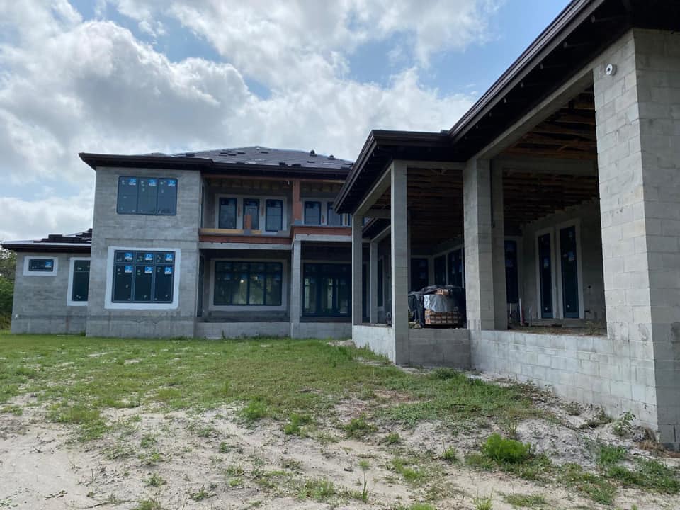 Two-story house under construction with gray cinder block walls, windows, and a green lawn.