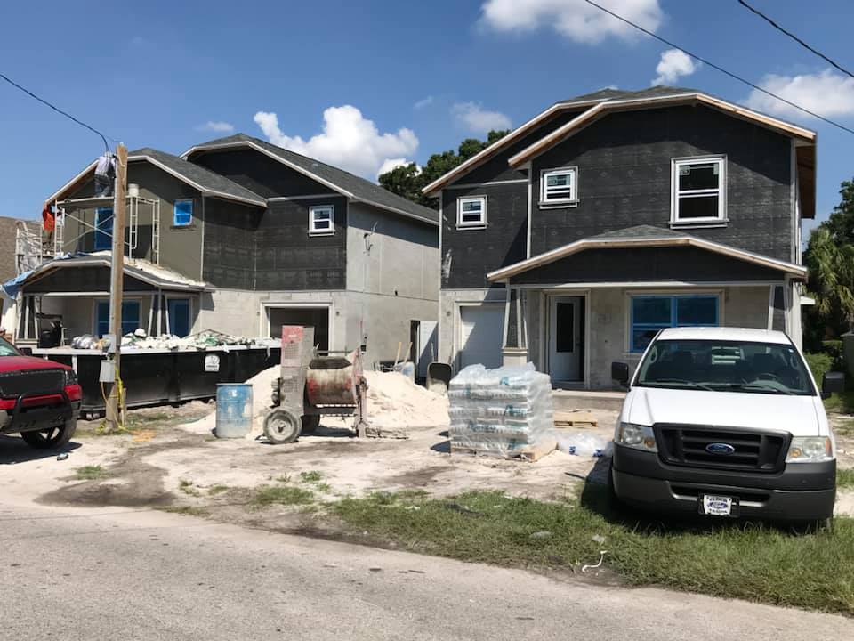 Two-story houses under construction, gray siding, blue window covers, construction equipment in front.