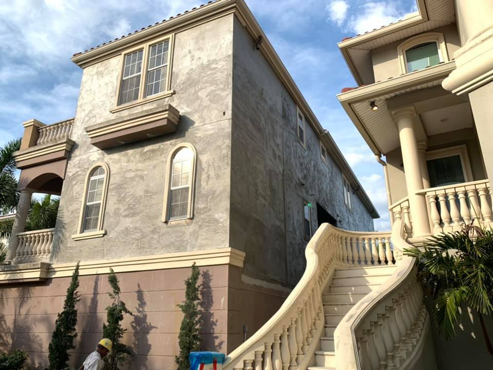 Two-story house under construction with stucco exterior, arched windows, and ornate staircase; sunny day.