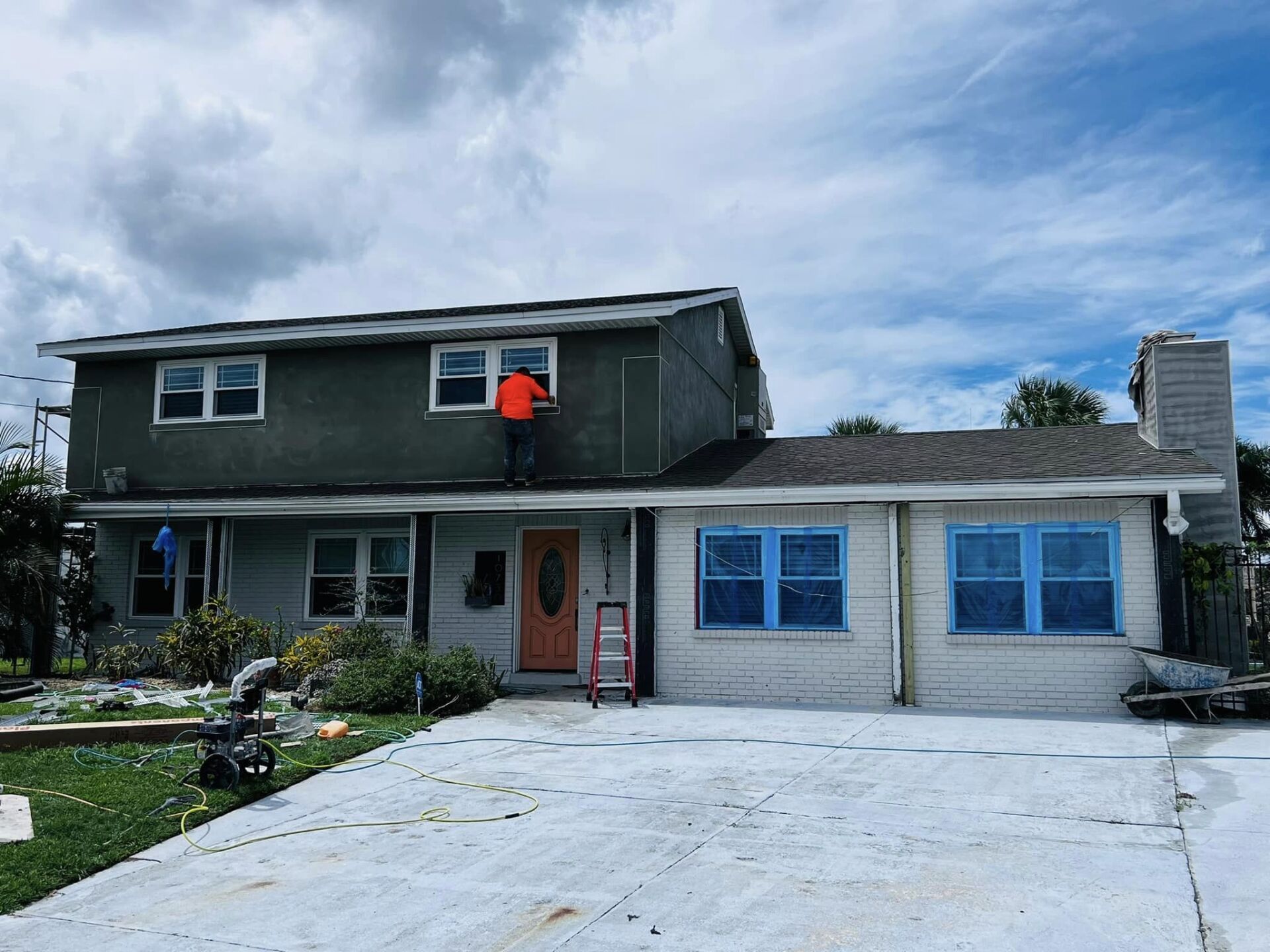 House under renovation with workers, some on ladders. Gray stucco, blue window covers.