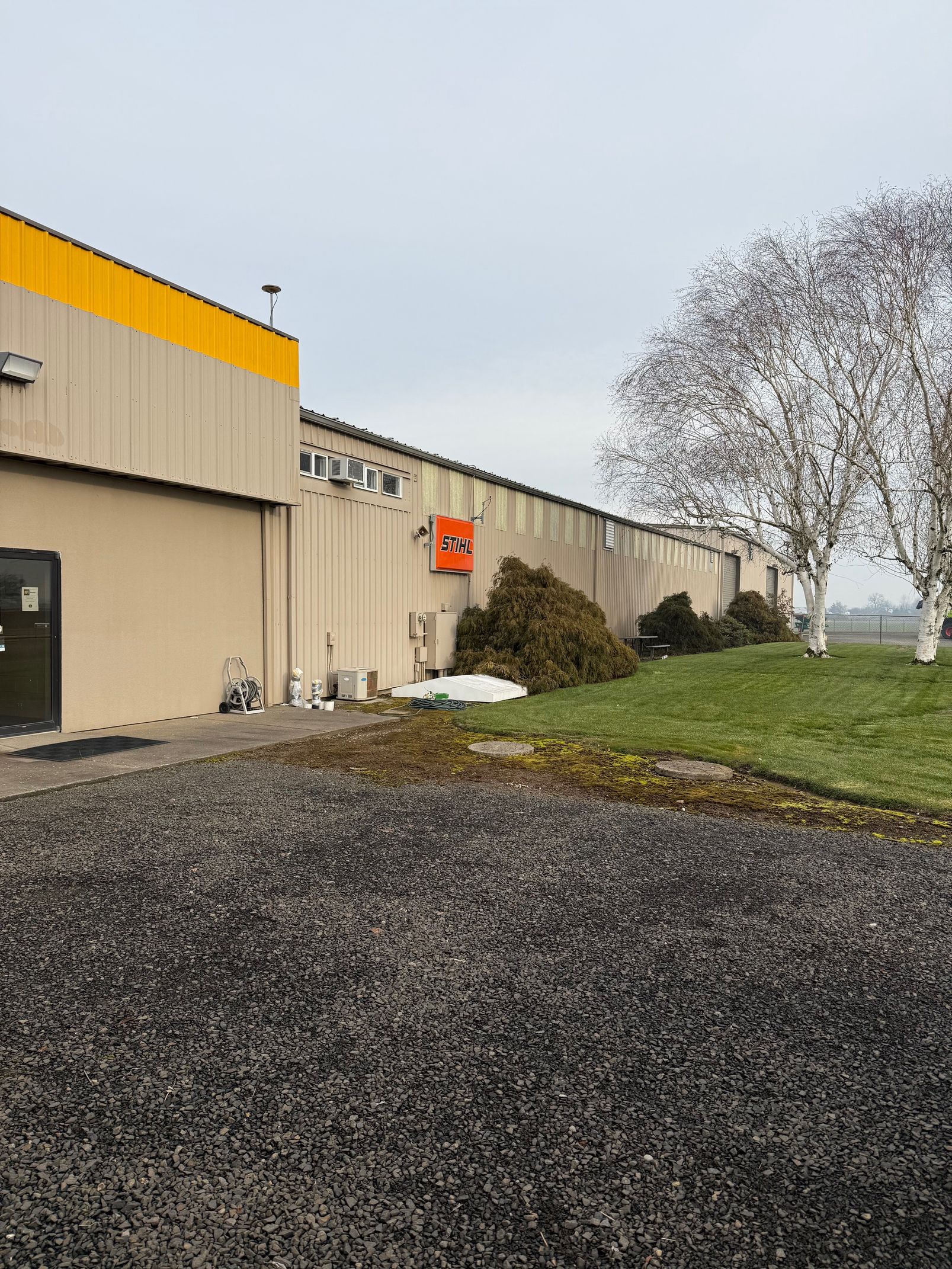 Industrial building with beige walls, yellow roof trim, gravel lot, and a grassy area by leafless trees under a gray sky