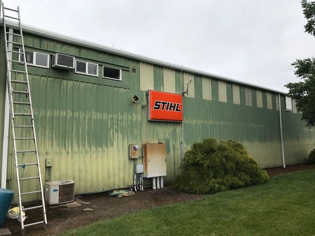 Green industrial building exterior with a STIHL sign, windows, a ladder, and shrubs by the wall.