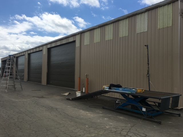 Industrial warehouse exterior with roll-up doors, a ladder, and a trailer under a cloudy sky