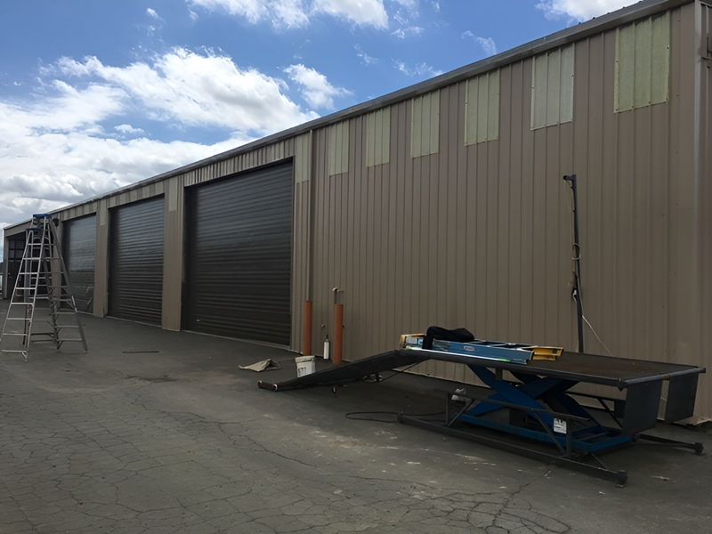 Industrial warehouse row with roll-up doors, blue sky, ladder, and a lift parked outside