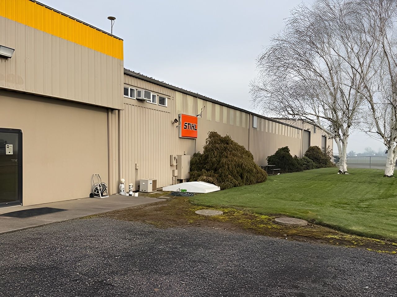 Beige industrial building with orange trim, a gravel driveway, and a leafless tree on a grassy lawn.