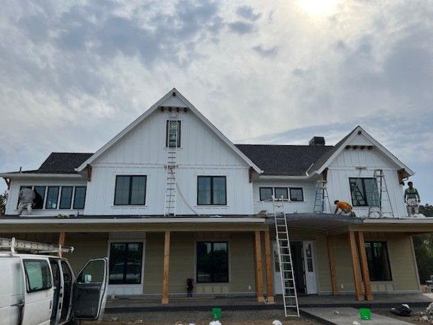 House exterior with white siding and gray roof under cloudy sky, ladder and van parked in front