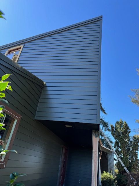 Gray-sided house with a steep upper wall and overhang against a bright blue sky.