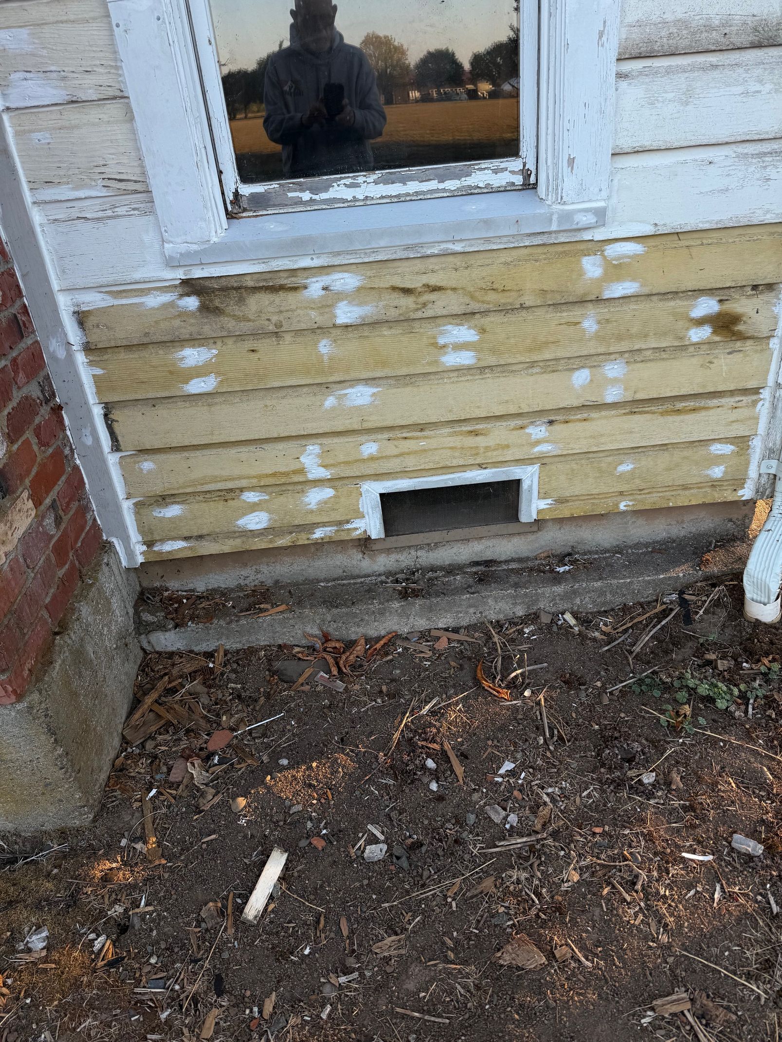 Peeling yellow house siding below a window with white trim and a small vent near the ground.