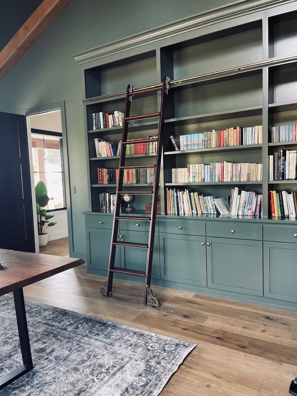 Bookshelves with a ladder in a teal-walled room beside a wooden table and rug