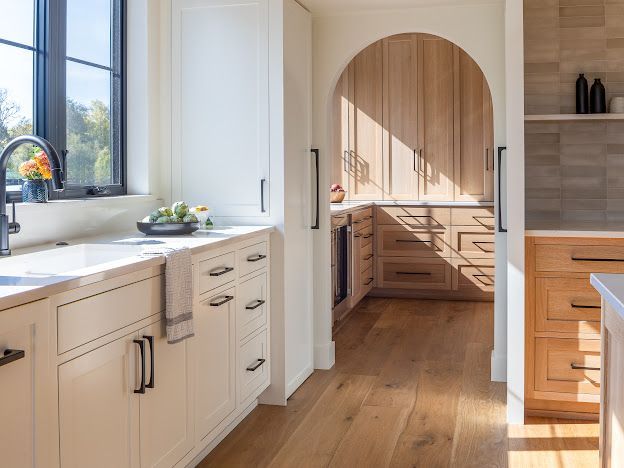 Bright modern kitchen with white cabinets, wood floors, and an arched doorway to a sunlit pantry