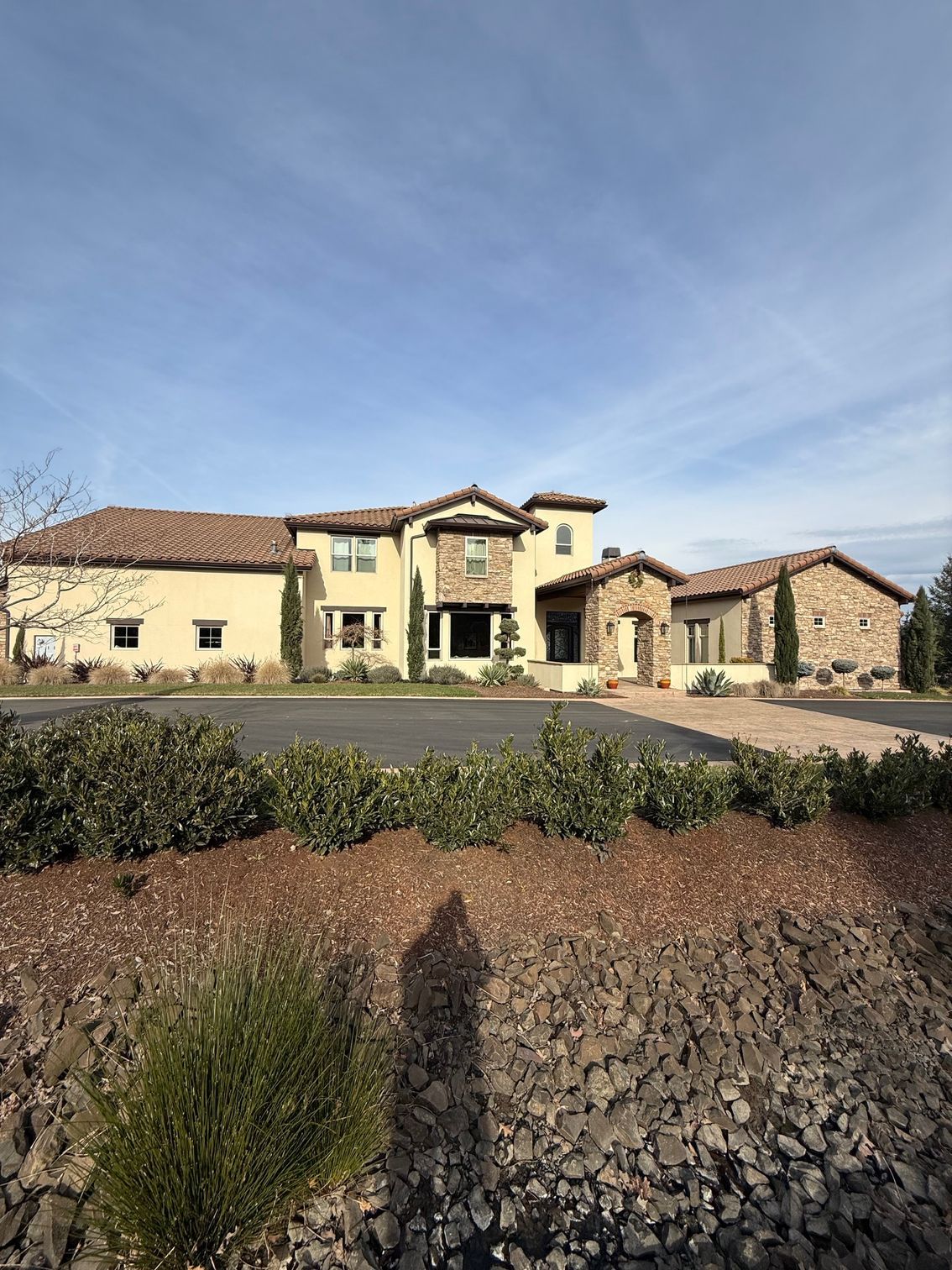 Large beige stucco house with red tile roof, viewed across a rocky landscaped foreground under a blue sky