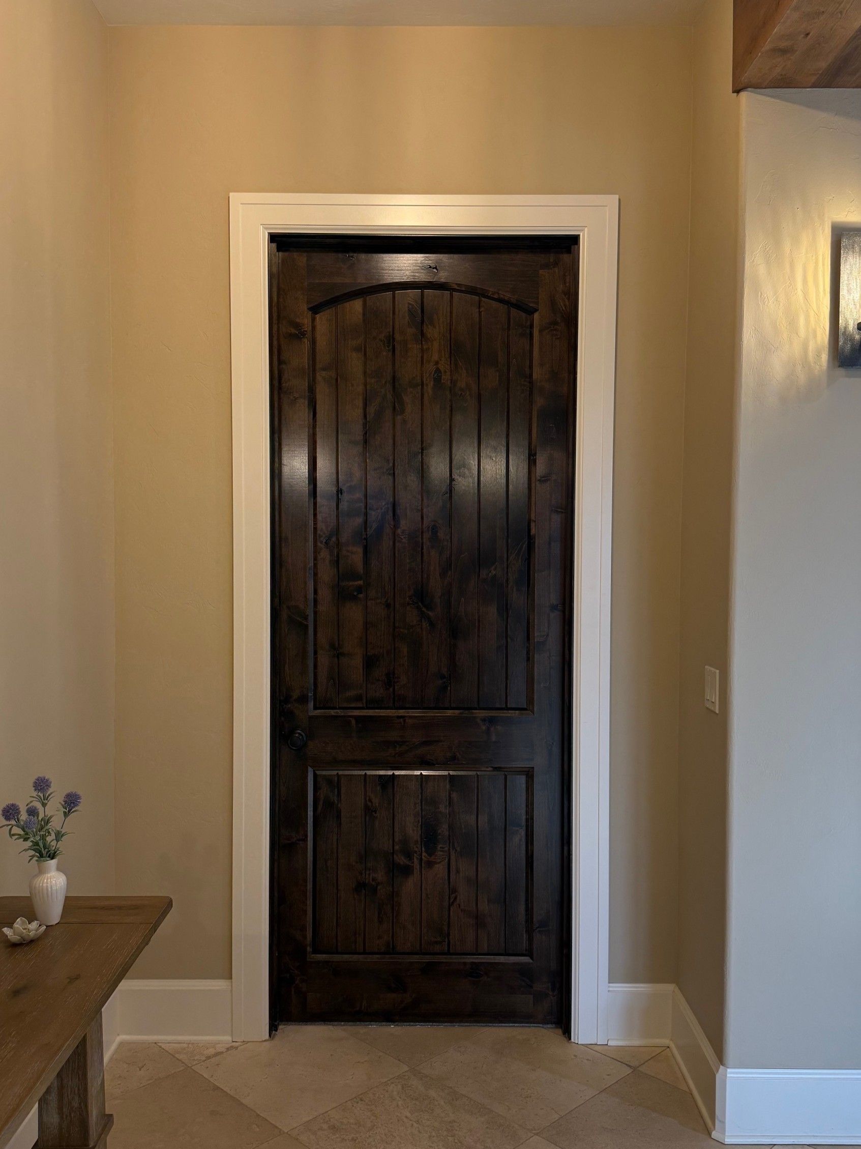Dark wood arched door in a beige hallway with white trim, a wall sconce, and a small table with flowers.