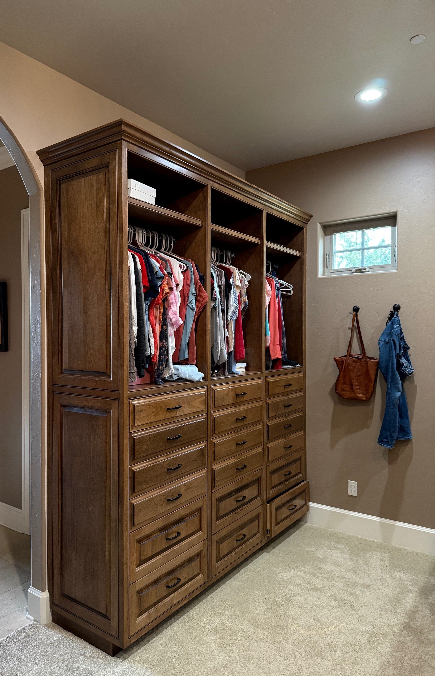 Wooden built-in wardrobe with hanging clothes and drawers in a beige bedroom/closet area.