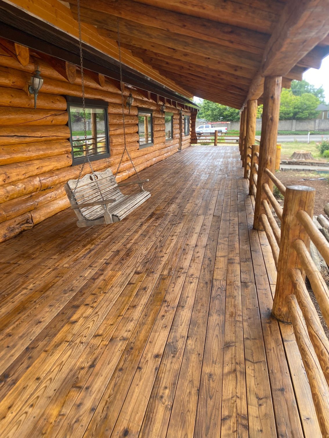 Wooden porch of a log cabin with wet floorboards, railing, windows, and a wooden bench chair.
