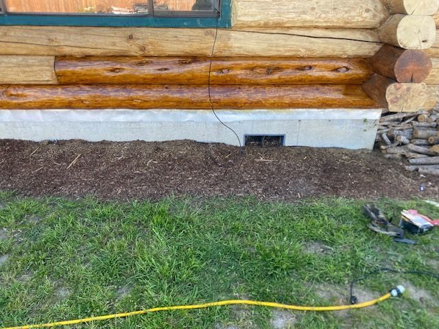 Log cabin wall with a small basement vent, mulch bed, grass, and a yellow hose in the foreground