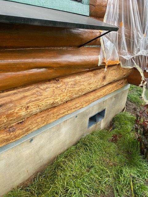 Close-up of damaged log siding and rotting wood beneath a house window, with torn plastic sheeting and grass below