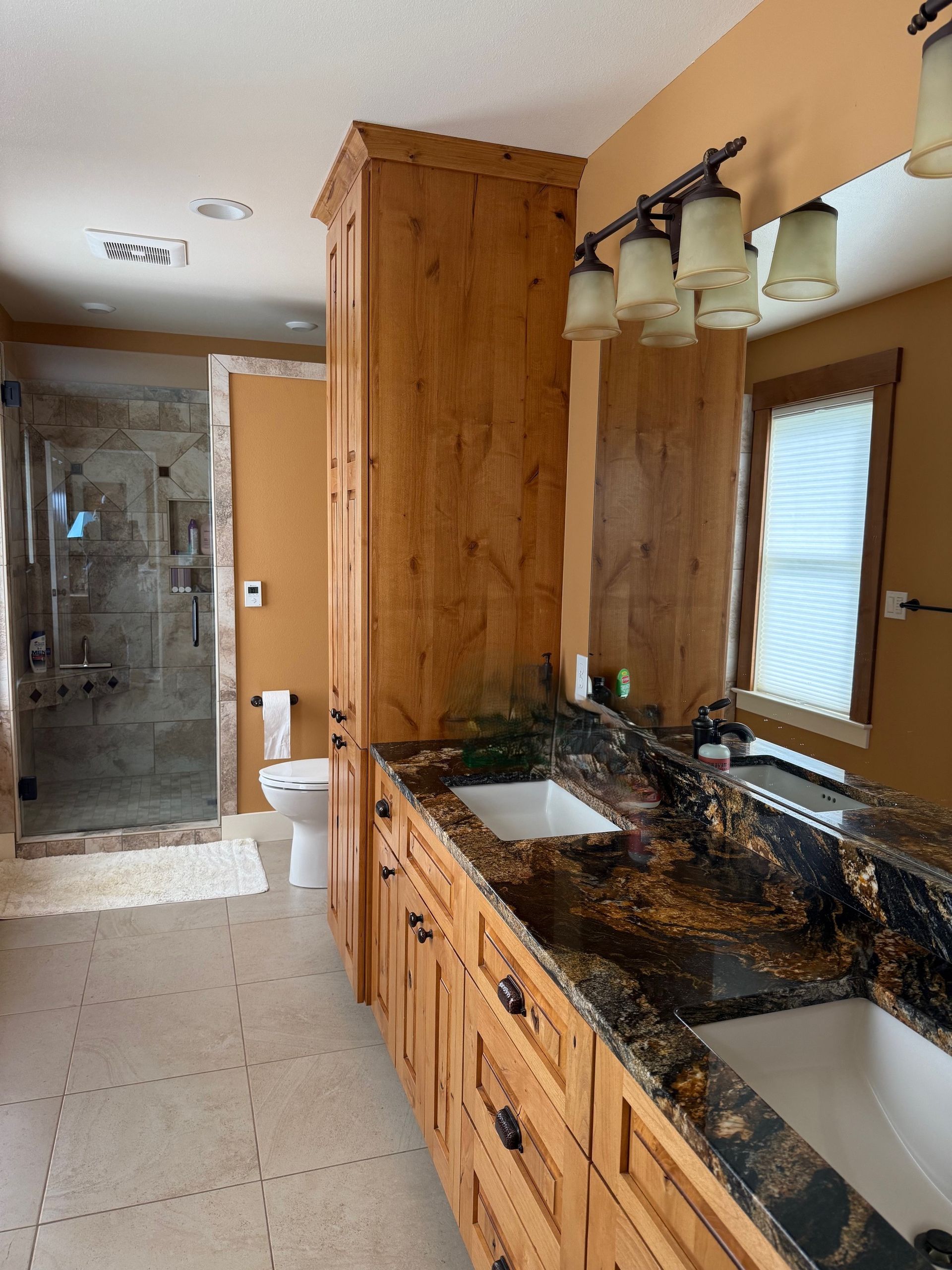 Bathroom with granite double vanity, wood cabinets, large mirror, window, and tiled shower.
