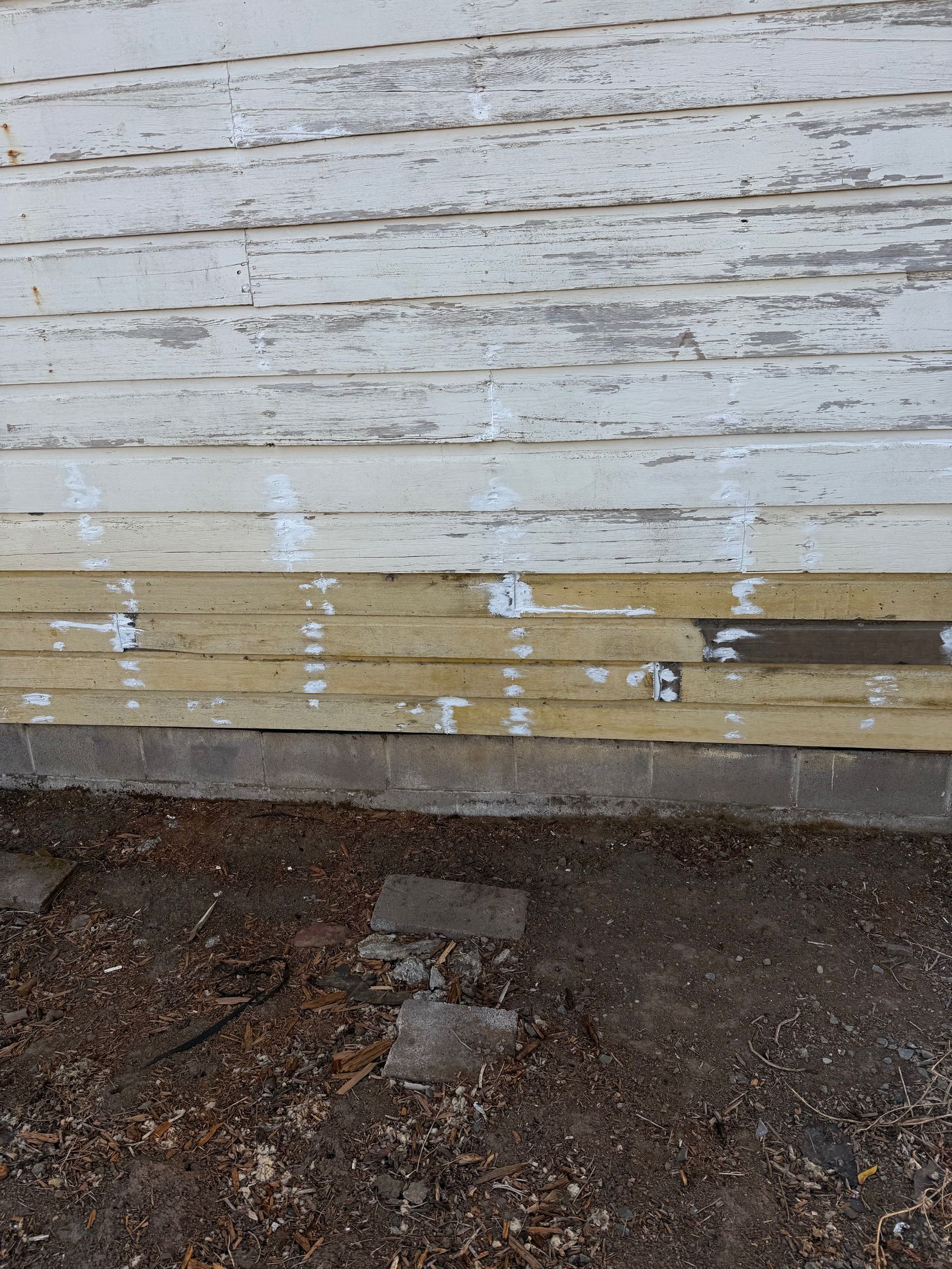 Weathered white wooden wall with peeling paint above a bare dirt strip and a small cinder block path