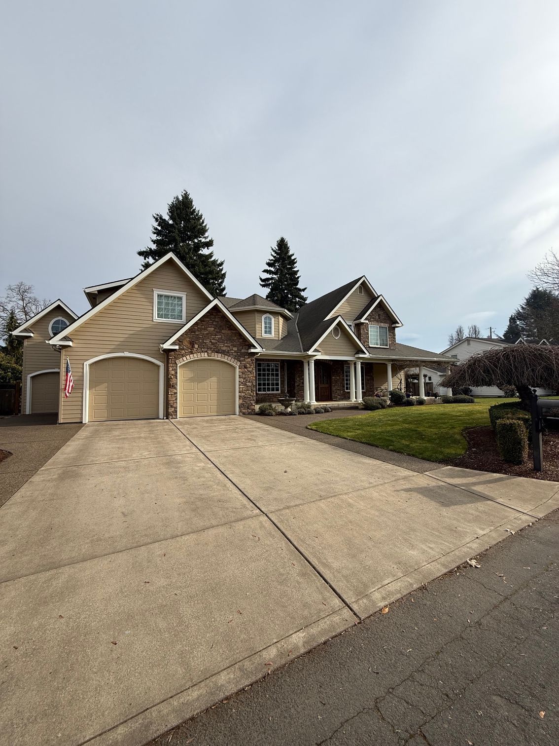 Suburban brick house with a wide driveway, front porch, and manicured lawn under a cloudy sky