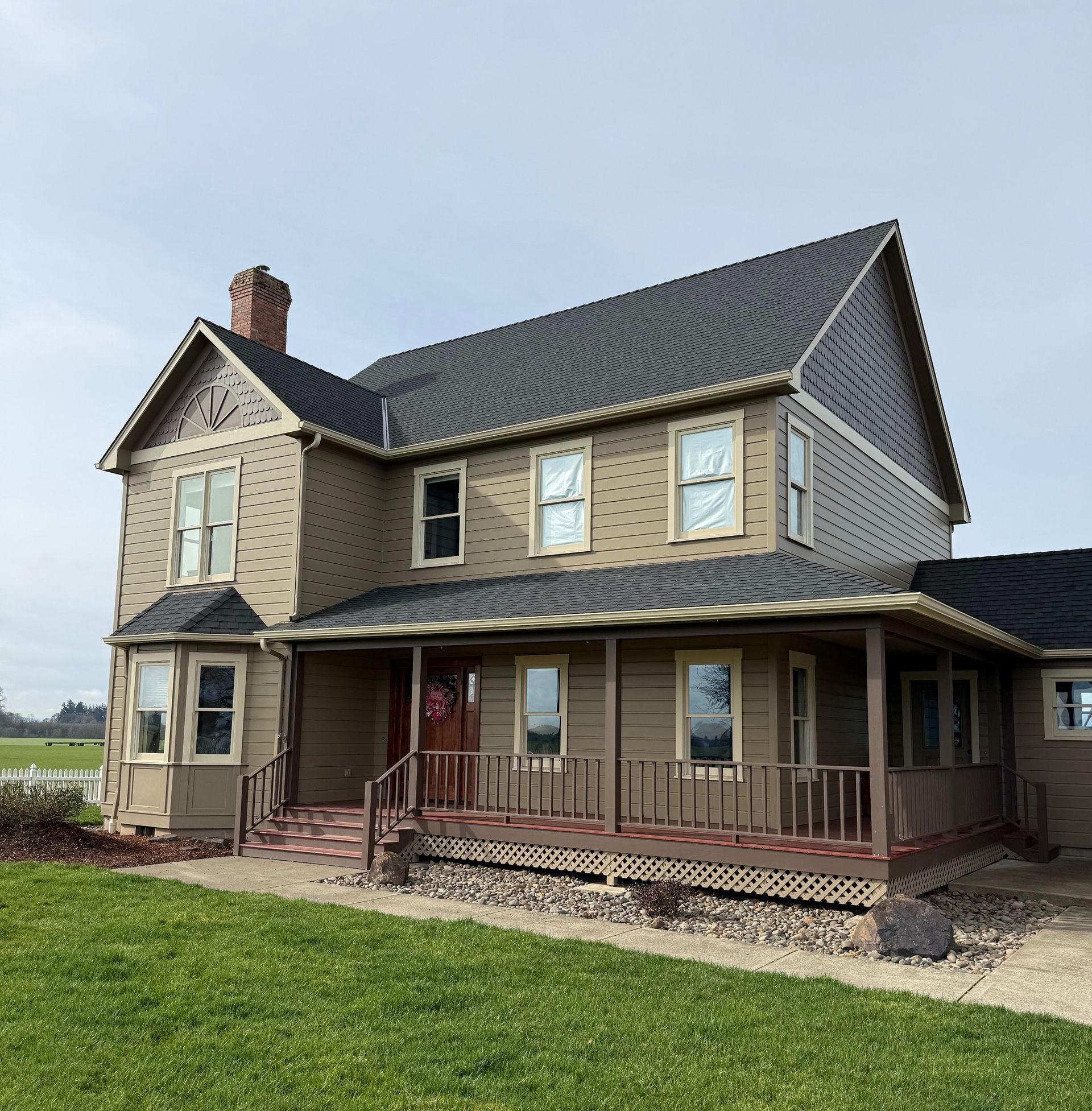 Two-story beige house with brown roof, front porch, and green lawn under a cloudy sky