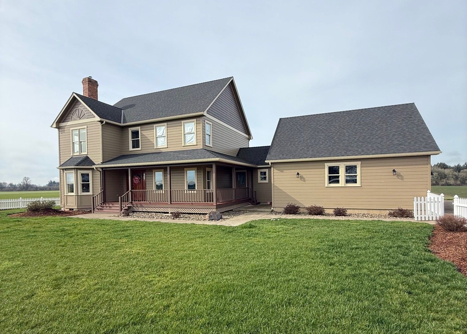 Two-story beige farmhouse with a covered porch and detached garage on a green lawn under a cloudy sky