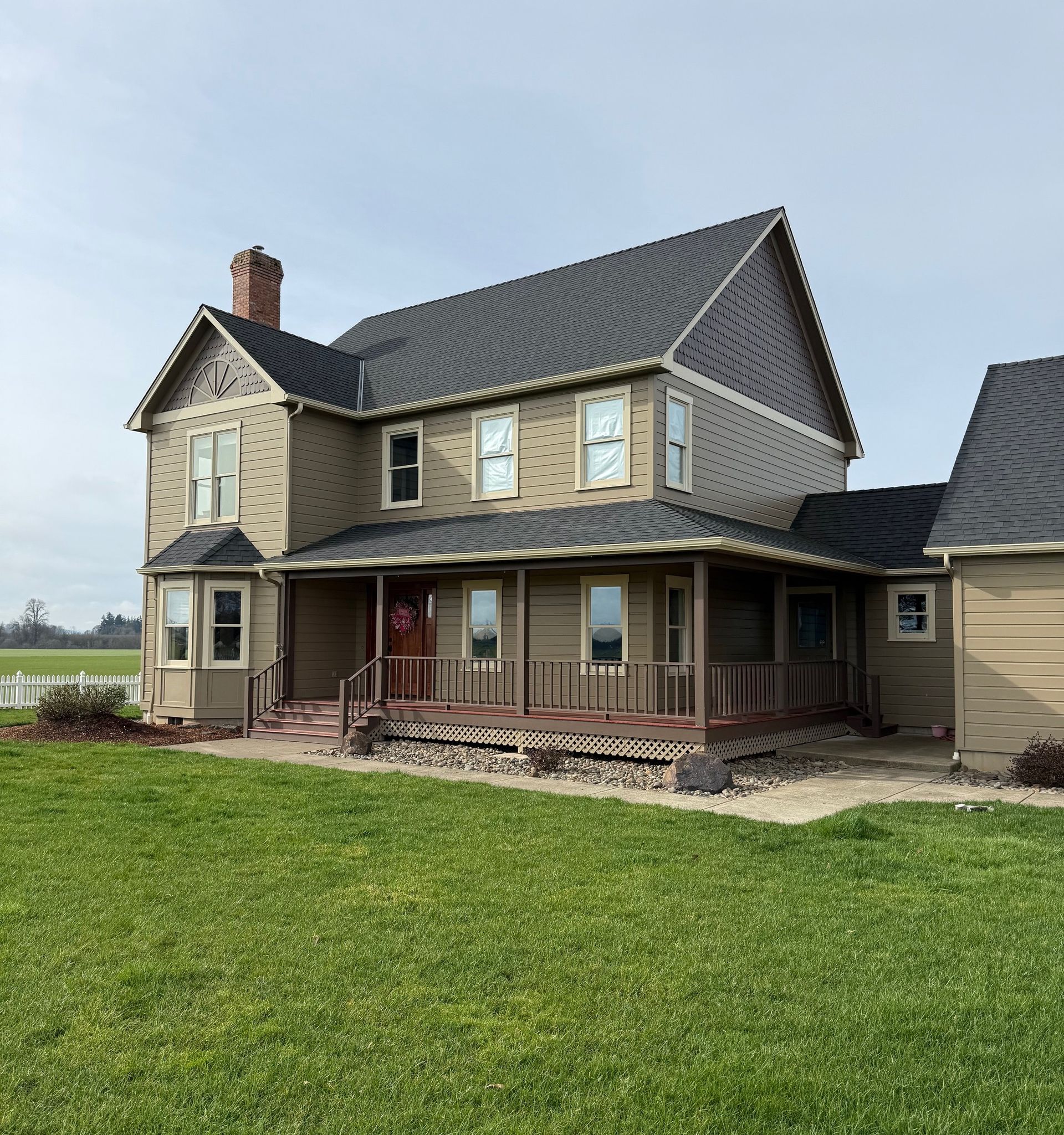 Two-story farmhouse with dark roof and covered porch beside a green lawn under a clear sky