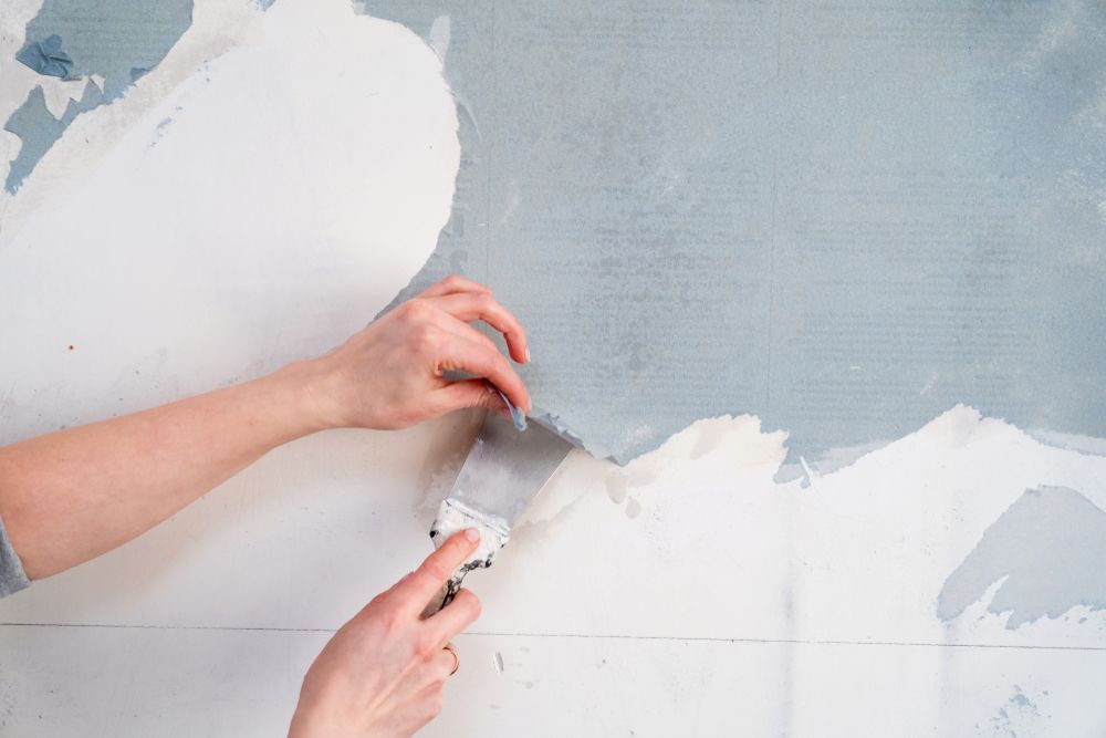 Hands scraping peeled paint from a light blue wall with a putty knife