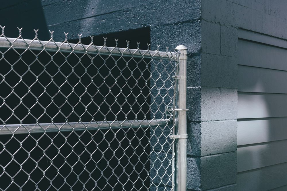 Chain-link fence beside a dark gray building with horizontal siding and a white gate post